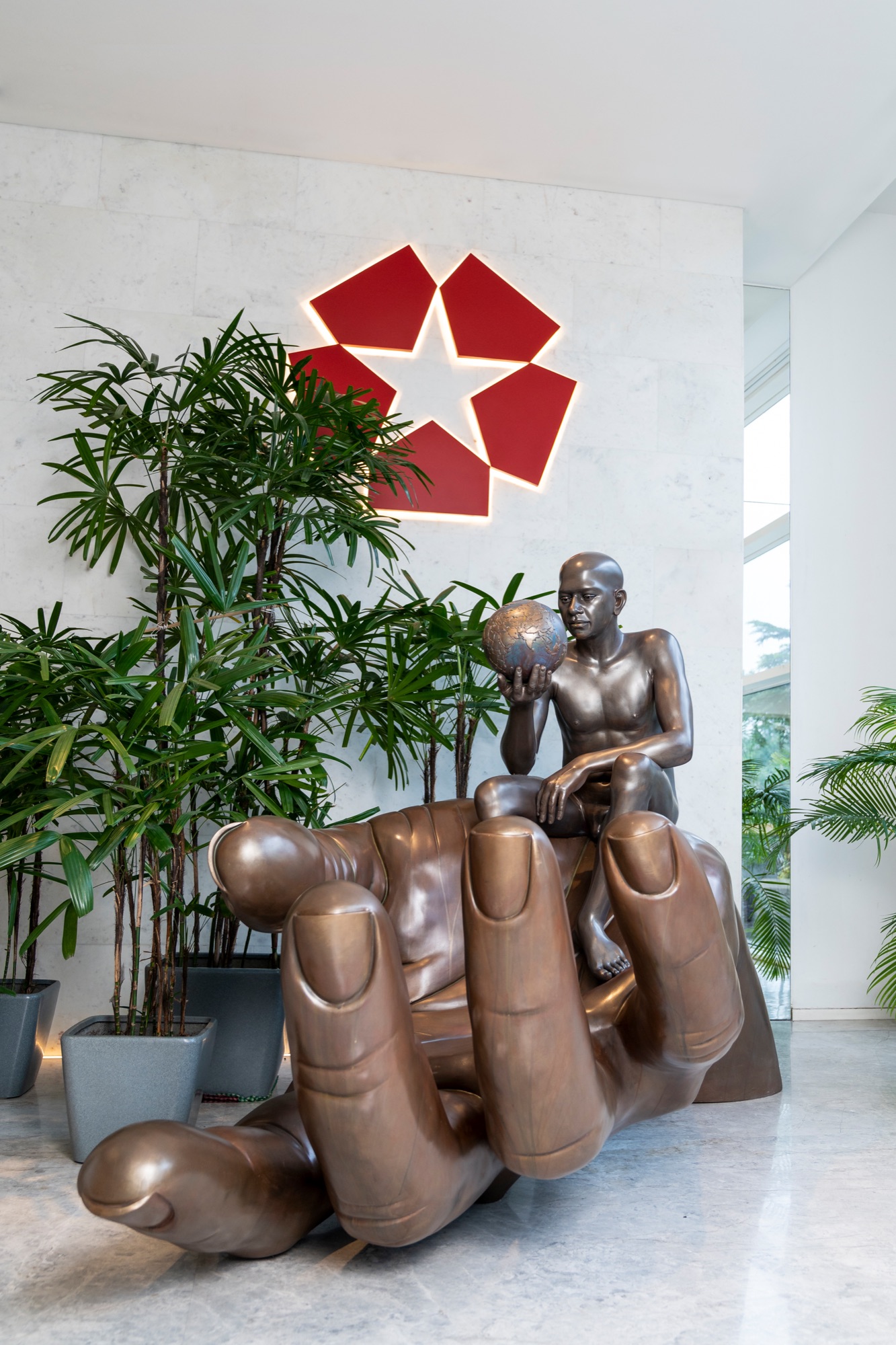 A corporate lobby with a bronze sculpture of a figure seated in a cupped hand beneath a red star wall relief — The Artemist, commercial